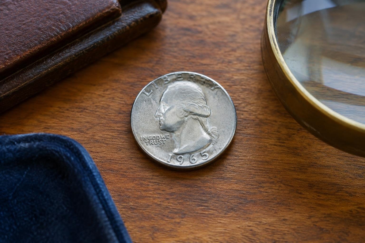 Clear obverse and reverse views of an ordinary 1965 Washington quarter A silver coin lies on a wooden surface between a leather case, a brass-rimmed magnifying glass, and a piece of dark blue fabric.